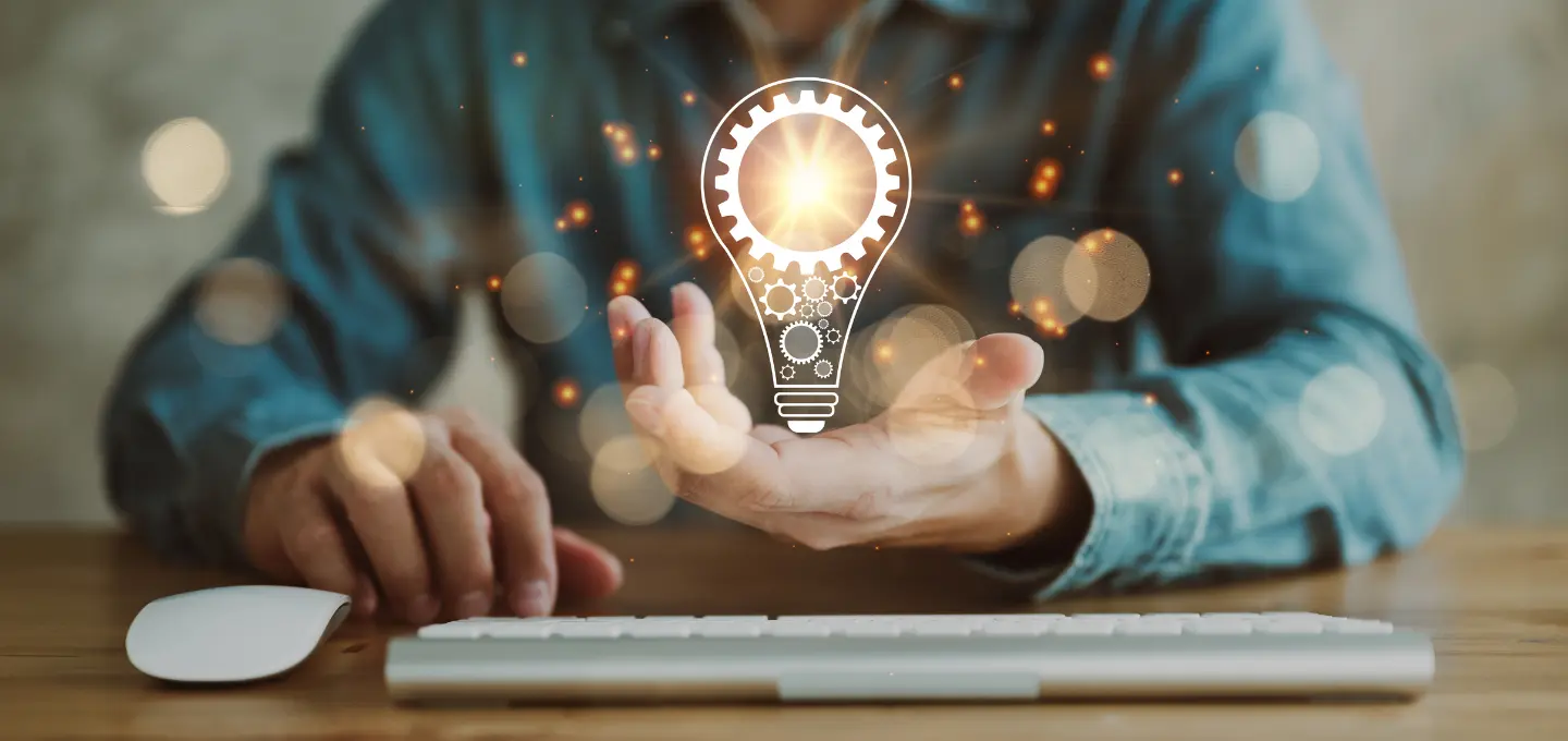 Photograph showing a person in a denim shirt using a keyboard and mouse with a glowing lightbulb icon featuring gears floating above an open hand. The scene symbolizes innovation, creativity, and technology in a work or digital environment, highlighted by warm lighting and bokeh effects.