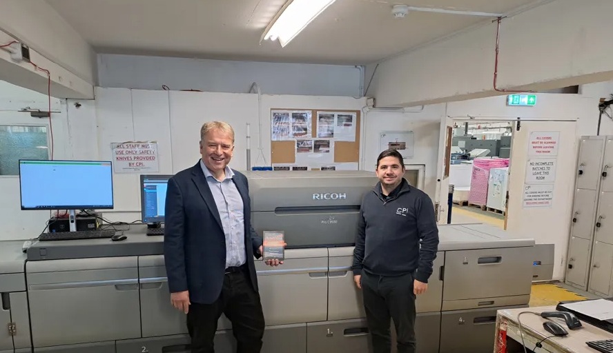 CPI Print - Twee mannen die glimlachen in een printfaciliteit, staand voor een grote Ricoh‑printer. Eén van hen houdt een geprint item vast. De ruimte is industrieel en georganiseerd.