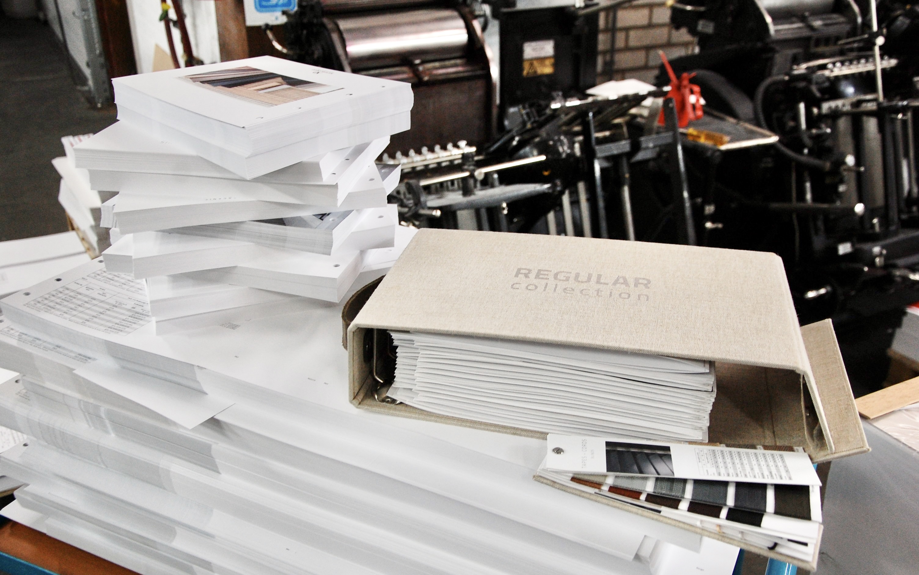 Photograph of a printing workshop showing stacks of printed materials and a beige binder labeled "REGULAR collection" filled with documents. The scene highlights production volume and organization, with printing machinery visible in the background and color swatches placed beside the binder.