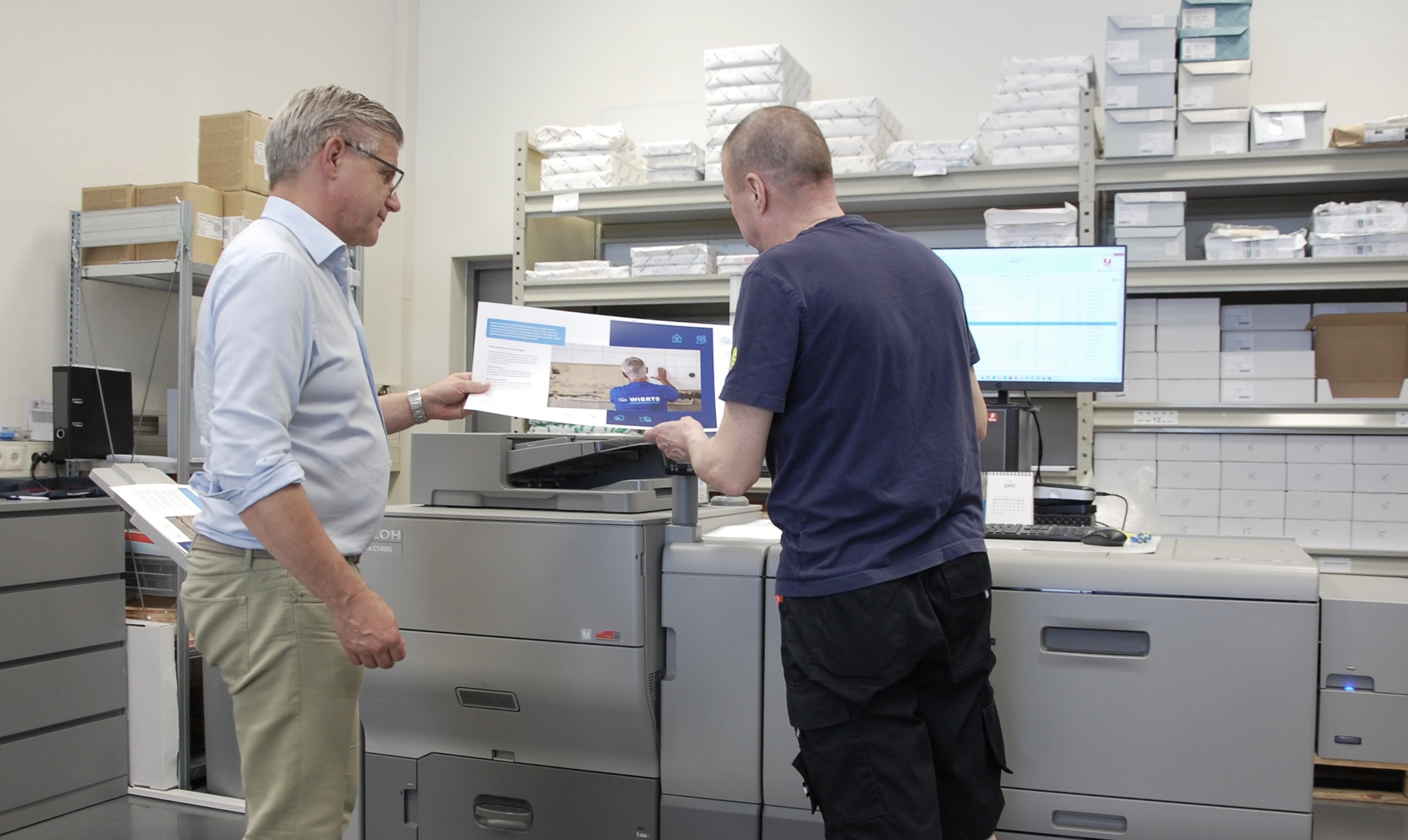 Twee medewerkers bekijken een geprinte brochure naast een productieprinter in een printomgeving.