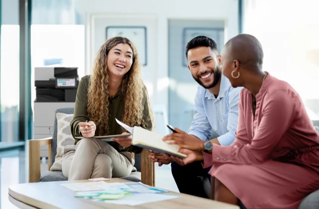 Three smiling young adults are sitting in a bright, modern office space, looking at papers and talking together. A Ricoh printer is visible in the background behind them.