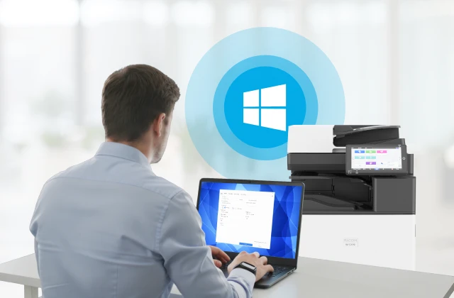 Man using a laptop next to an office printer, with a Windows icon indicating wireless printing setup.