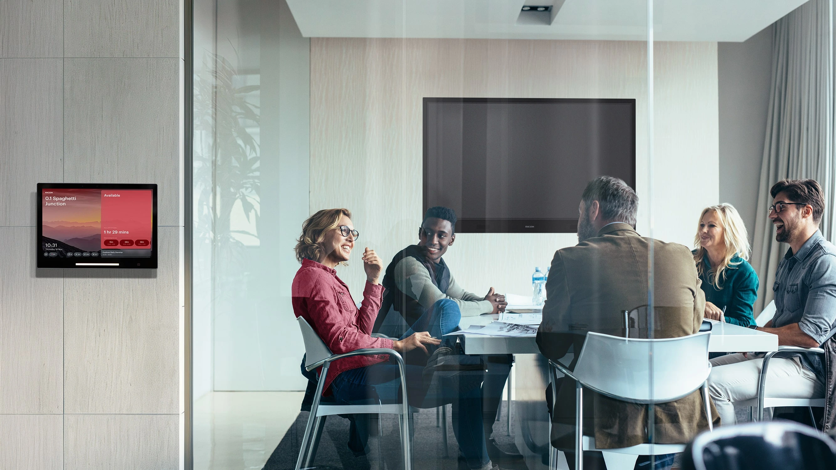 5 mensen zitten in een vergaderruimte, het vergaderruimtebedieninspaneel is zichtbaar aan de buitenkant van de meetingroom