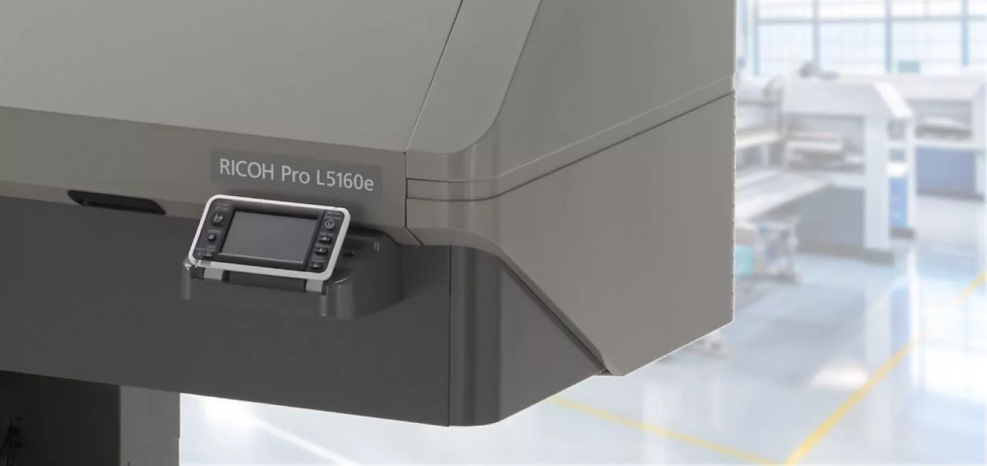  A close-up of the control panel on a large industrial printer, specifically a "RICOH Pro L5160e". The touch screen is visible, but the display is off. The background is a blurry shot of a print shop with other machinery.