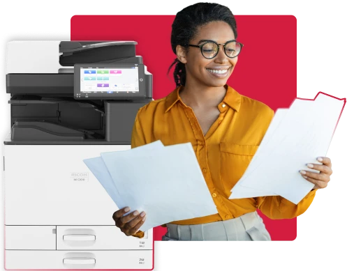 A smiling woman in an orange shirt and glasses holds stacks of white papers, standing in front of a white Ricoh printer.