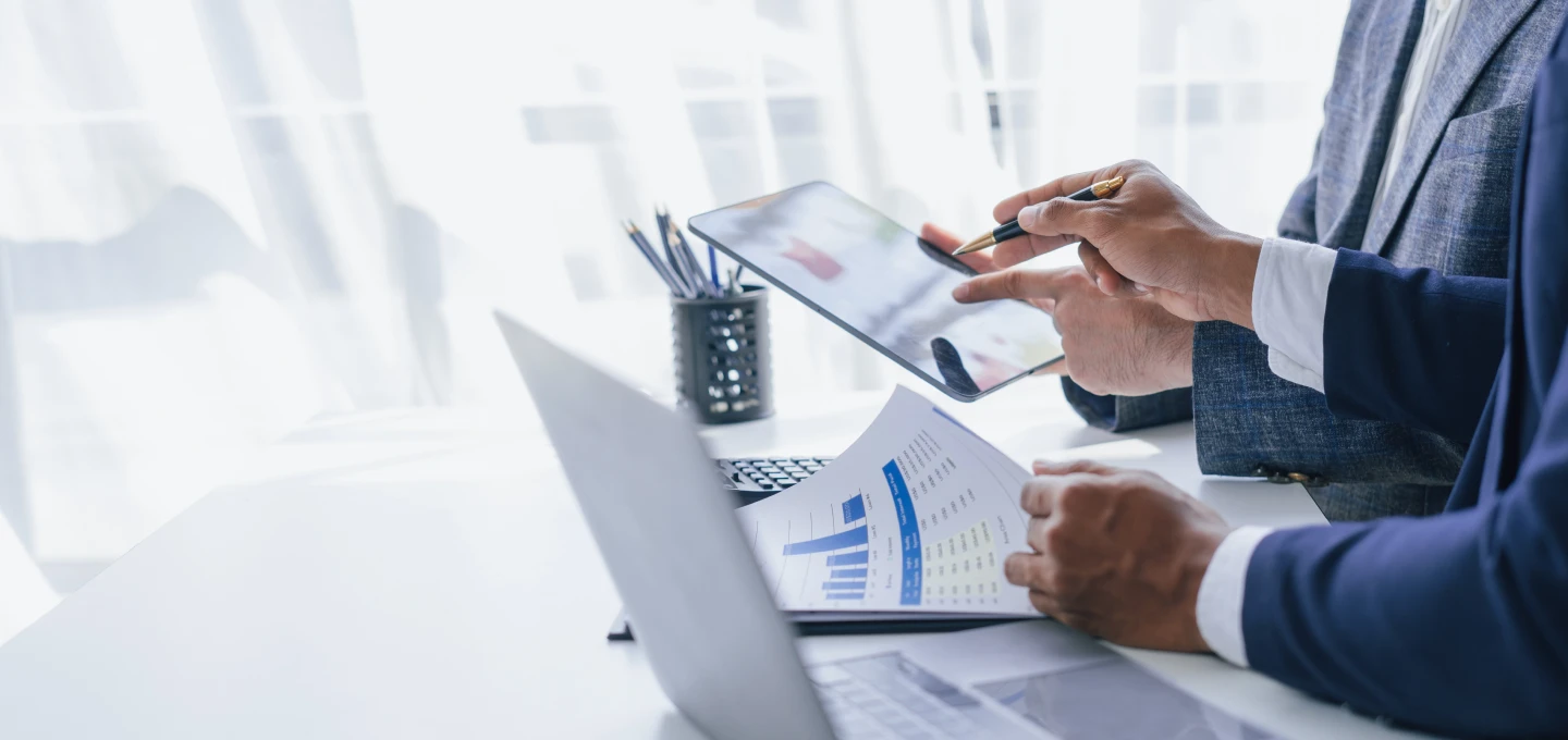 Two people in business attire are reviewing data on a tablet and a laptop. One person is pointing at the tablet screen, while the other is looking at a document with graphs.