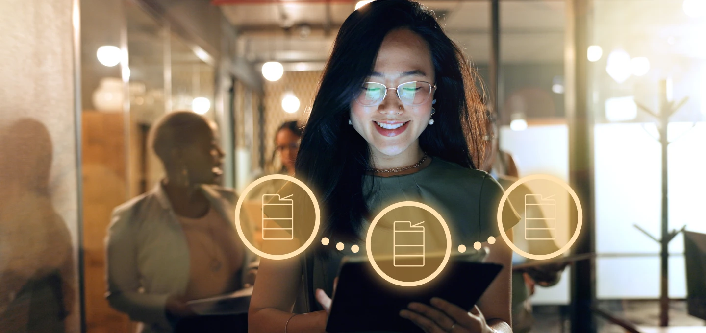 A person with long dark hair and glasses smiles while looking down at a tablet they are holding. Their face is illuminated by a glow, possibly from the screen. Three glowing circular icons, each containing an image of a printer, float in front of them. In the background, other blurred figures are visible in what appears to be an office hallway.