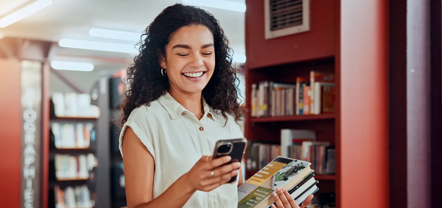 Photograph of a person standing in a library holding a smartphone in one hand and several books in the other. Background includes bookshelves filled with books and soft lighting, suggesting a quiet, studious environment.